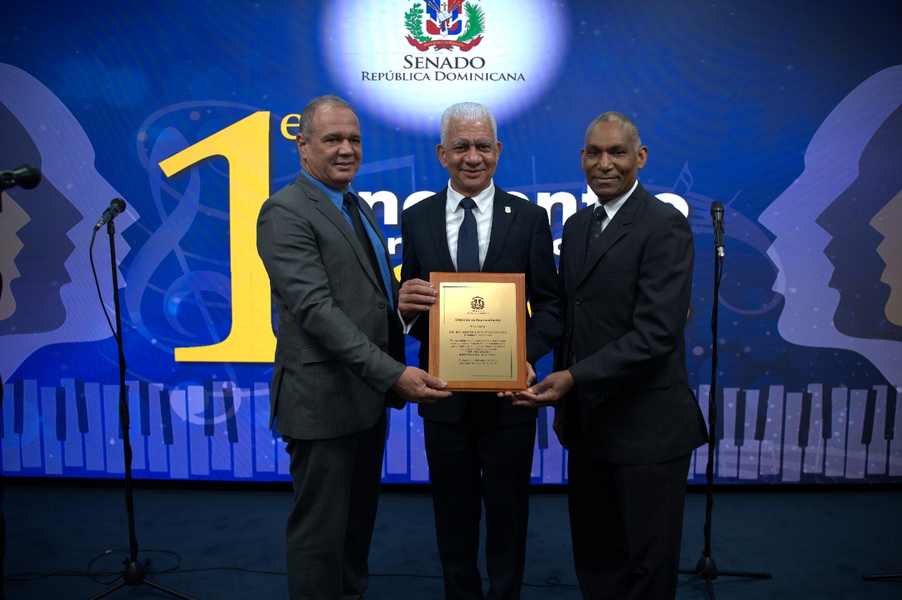 SANTO DOMINGO. – El Senado de la República, a través de la Dirección de Representación, celebró el Primer Encuentro Interinstitucional de Coros, una iniciativa que reunió a agrupaciones del Conservatorio Nacional de Música, el Virgen del Amparo (Armada Dominicana), San Miguel Arcángel (Ejército Nacional), Nuestra Señora del Carmen (Fuerza Aérea), el coro del Poder Judicial, San Judas Tadeo (Policía Nacional) y el coro del Senado de la República. La jornada destacó la diversidad y riqueza vocal proveniente de distintas instituciones del Estado. Durante el evento, el presidente de la Cámara Alta, Ricardo de los Santos, propuso coordinar con el Ministerio de Cultura la institucionalización y expansión nacional de este tipo de encuentros, resaltando la calidad interpretativa exhibida. “Es algo que debemos repetir”, afirmó tras recibir un reconocimiento por su respaldo al fortalecimiento del coro institucional, destacando el impacto cultural de la actividad. El director del Departamento de Representación, Leonardo Peña Javier, valoró la trascendencia histórica de este primer acercamiento coral, al manifestar expresiones del folclore dominicano y unir voces de distintas entidades estatales. “Más que un encuentro, este evento es un instrumento visual, artístico y cultural para la historia”, expresó, subrayando la importancia de promover espacios de identidad y creatividad. En el marco del acto celebrado en el Auditorio Eduardo Estrella, cada coro interpretó dos o tres piezas y recibió un reconocimiento de parte de autoridades legislativas y militares. Entre los galardonados estuvieron Edwin Disla, Armando Morillo, Germán Cornielle, Andrés Capellán, Elioenai Medina, Teodoro Reyes, así como Paola Suazo y Nelson Guzmán Diplán, figuras clave en el desarrollo del coro del Senado. A la ceremonia asistieron directores, funcionarios y colaboradores del Senado de la República, reforzando el compromiso institucional con la promoción cultural.